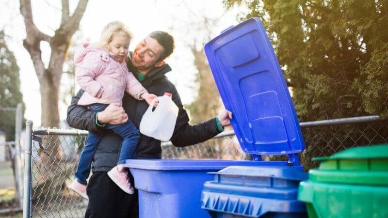Dad and daughter taking milk bottle out to the outside trash can.