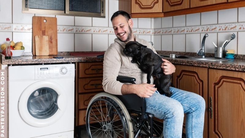 Man in wheelchair in kitchen with his dog on his lap.